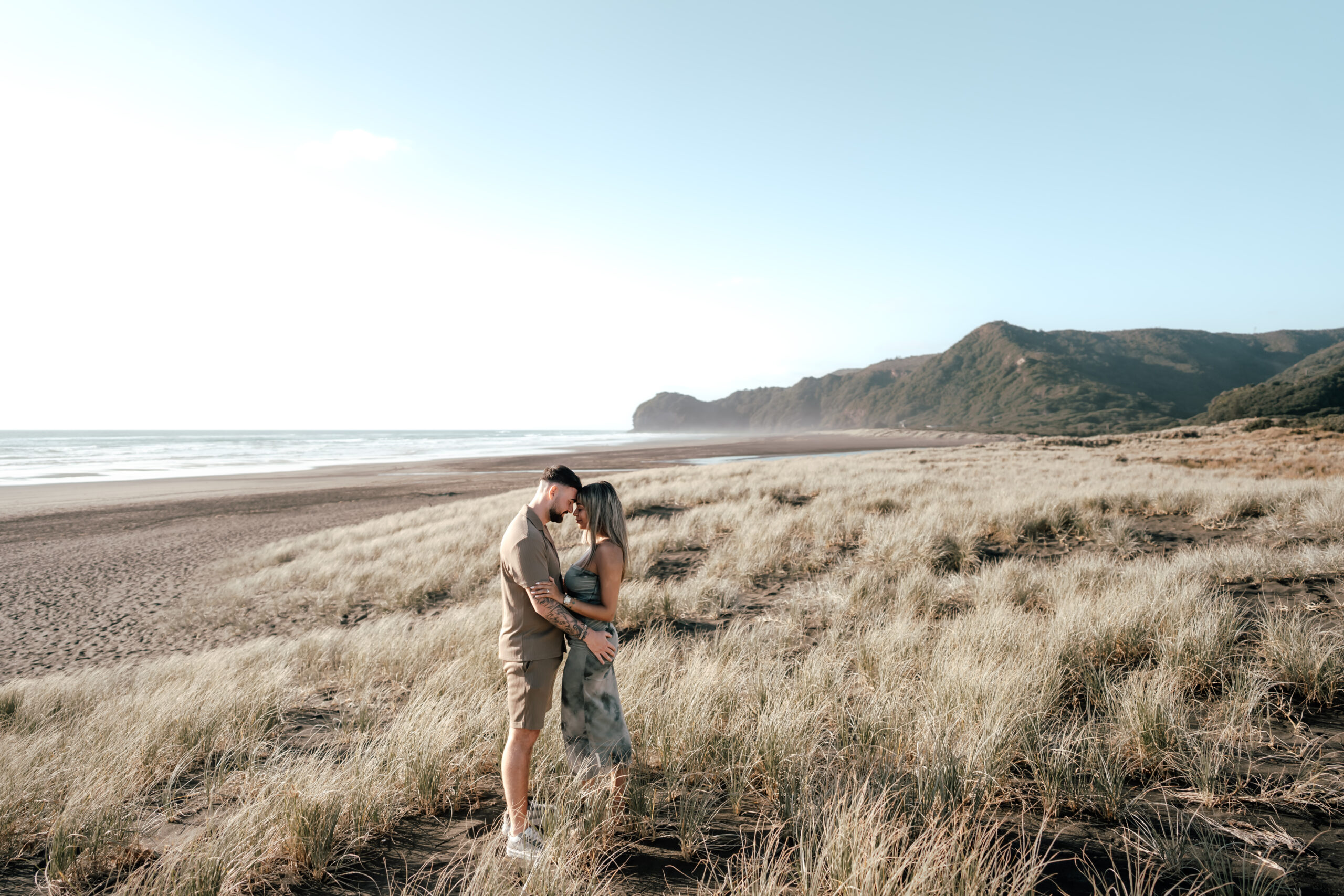 The couple standing with their heads together and the beautiful piha beach and sand dunes behind them during their proposal photoshoot