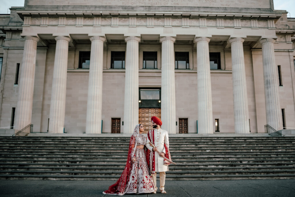 Indian couple in red and beige traditional wedding outfits during post-wedding photoshoot at Auckland Museum with grand columns and romantic poses