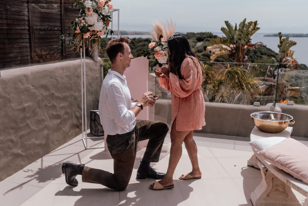 Romantic ocean view proposal at Delamore Lodge on Waiheke Island, with groom-to-be kneeling on a private balcony as his partner reacts with joy.