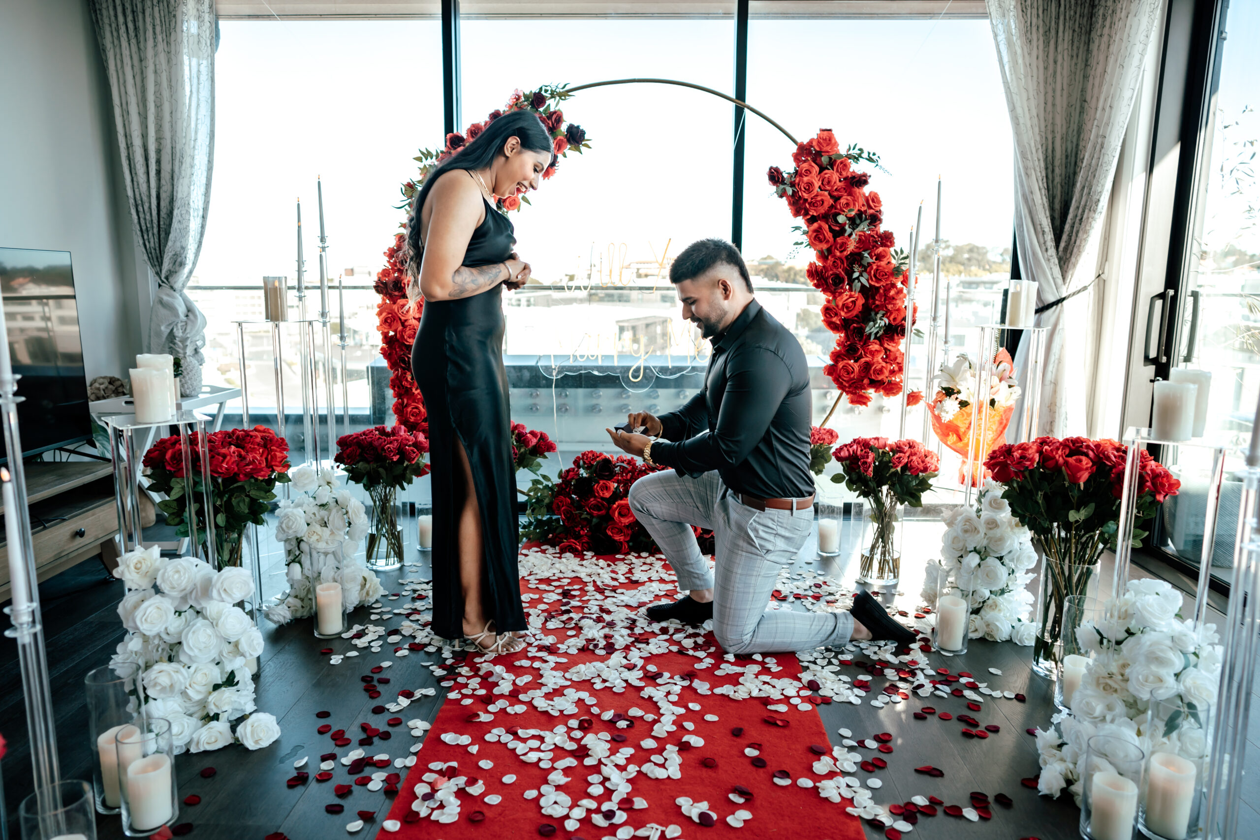 Romantic penthouse proposal in Auckland with red roses, candlelight, and floral backdrop as Daljit proposes to Jasleen surrounded by friends.