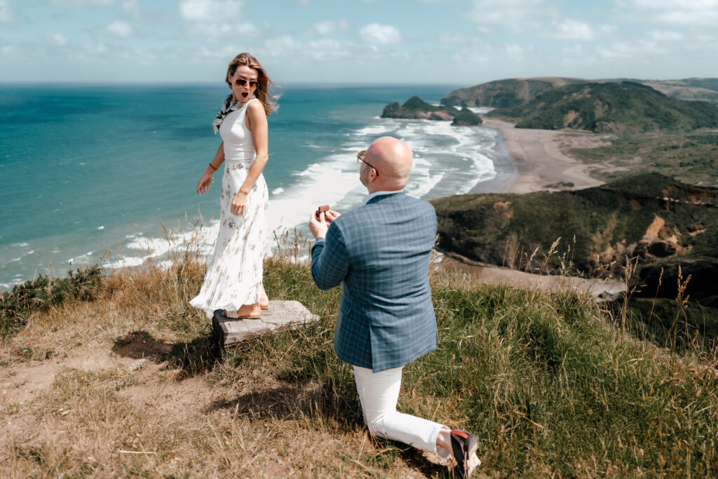 John proposing to Grace on a cliffside rock at Bethells Beach during a surprise helicopter proposal
