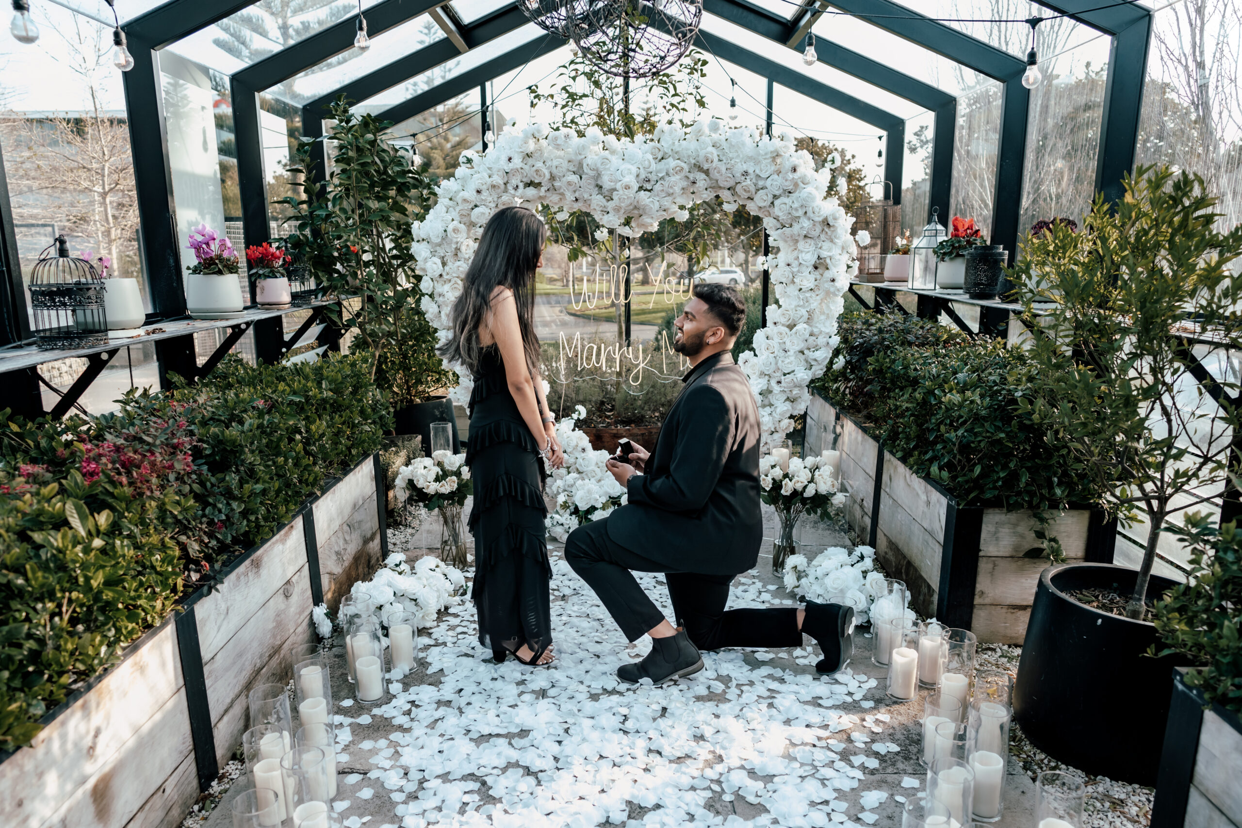 Elegant white rose proposal setup at The Grange in Takapuna, with heart-shaped floral arrangement, candles, and “Will You Marry Me” sign for Sanjesh and Niyanta’s surprise engagement