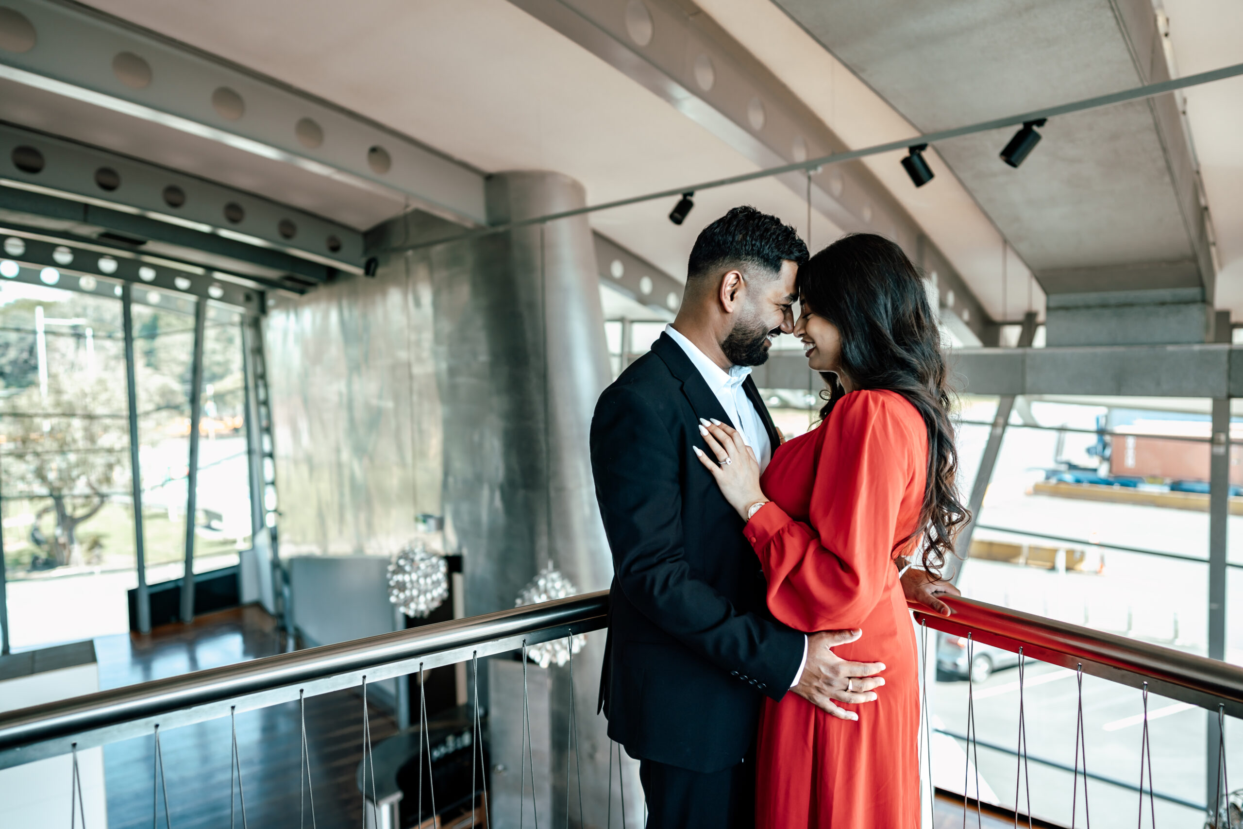 Romantic waterfront proposal at Mechanics Bay in Auckland featuring illuminated “Marry Me” letters, with Shivam in a tuxedo and Kyra in a red dress.