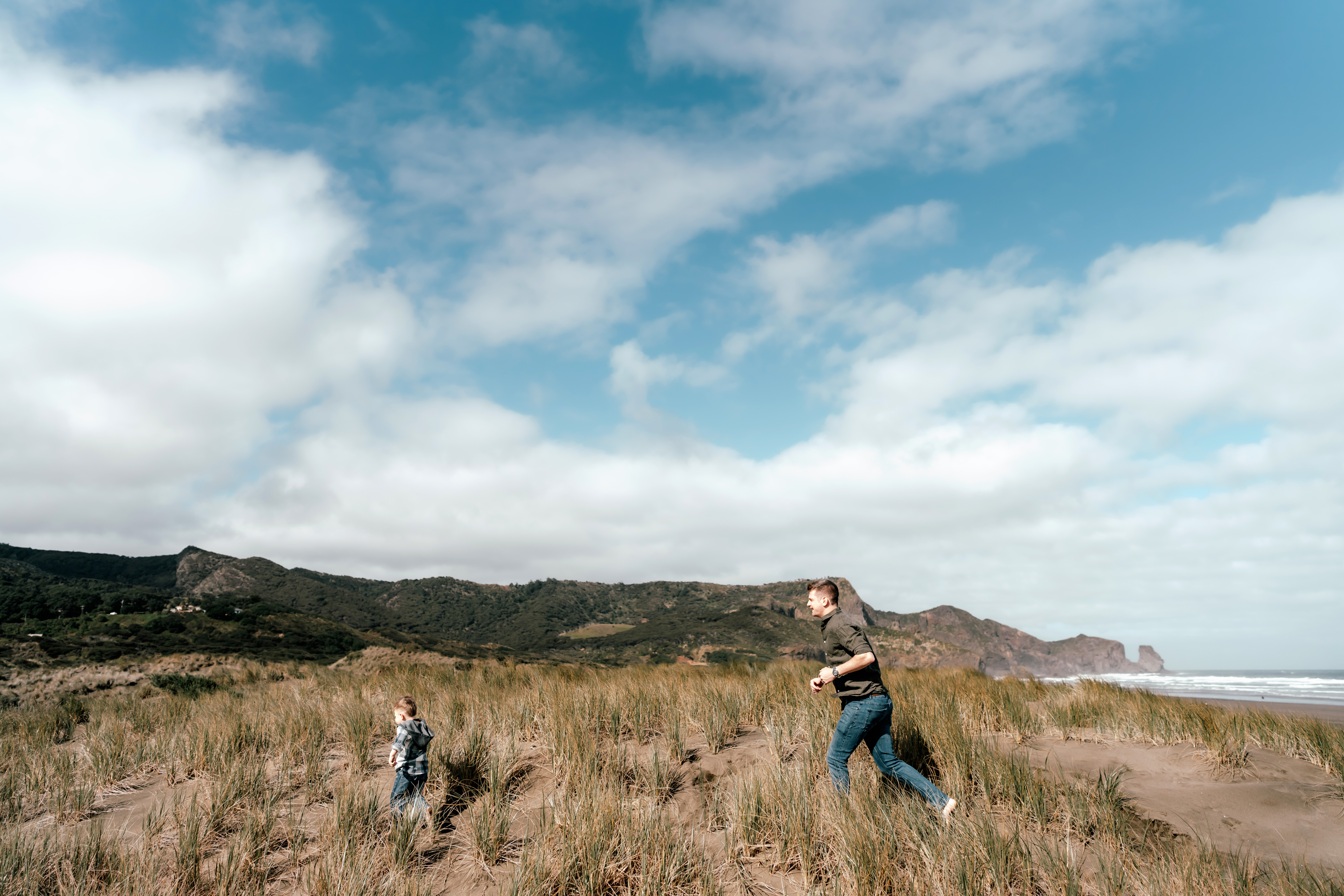 Family photoshoot at Bethells Beach in Auckland featuring dad and son running on the sand dunes with ocean and cliffs in the background.