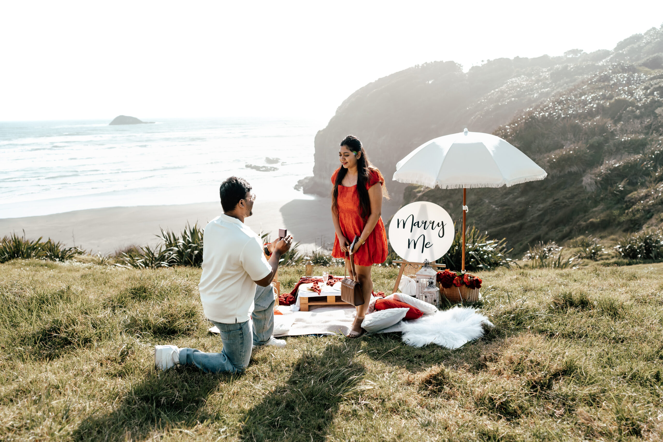 Dinesh proposing to Madhu on the clifftop overlooking Maukatia Bay Beach in Auckland during a romantic surprise proposal captured by Zanthe Vorsatz Photography