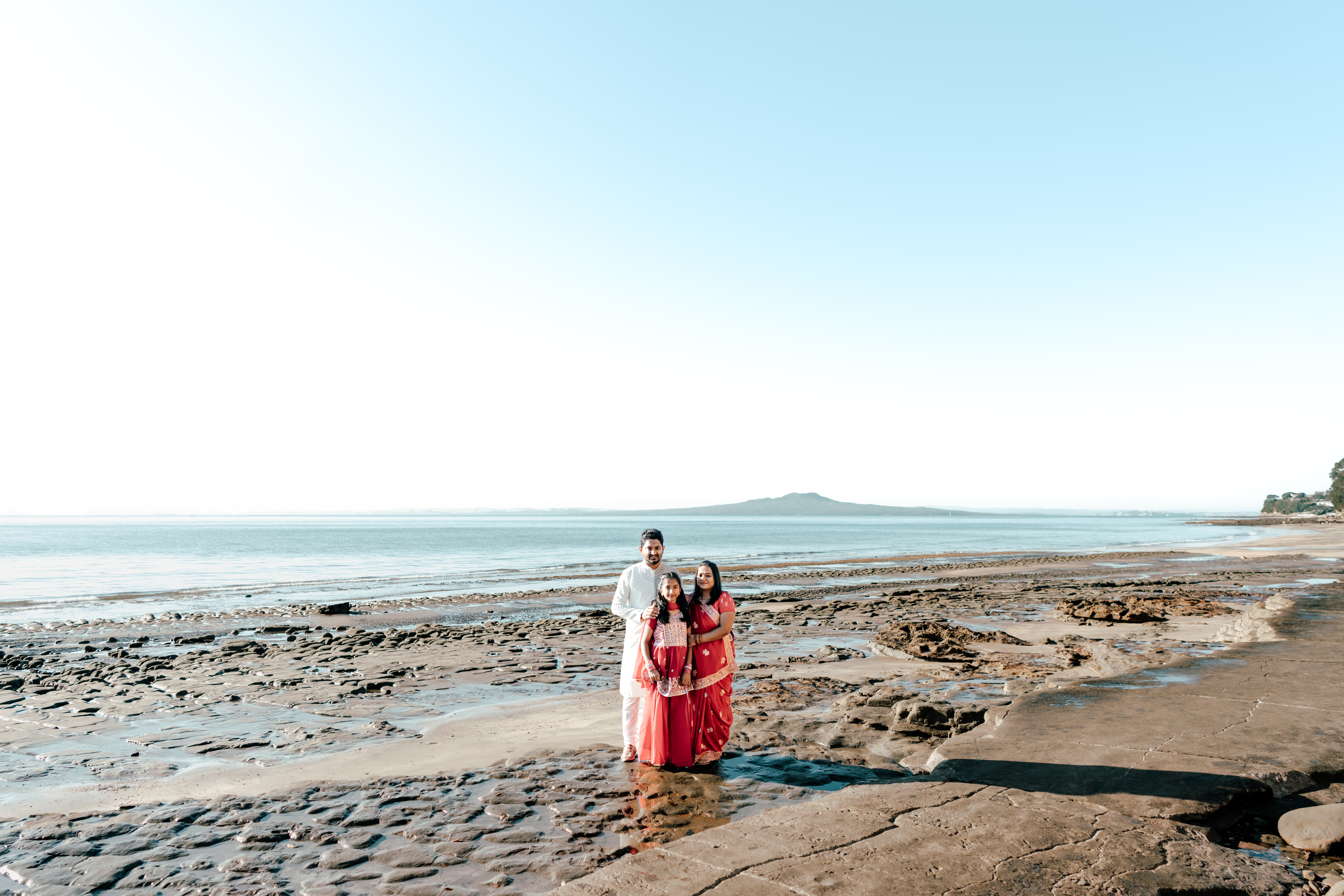 The Prasad family in vibrant traditional Indian outfits during a sunny beach family photoshoot in Auckland