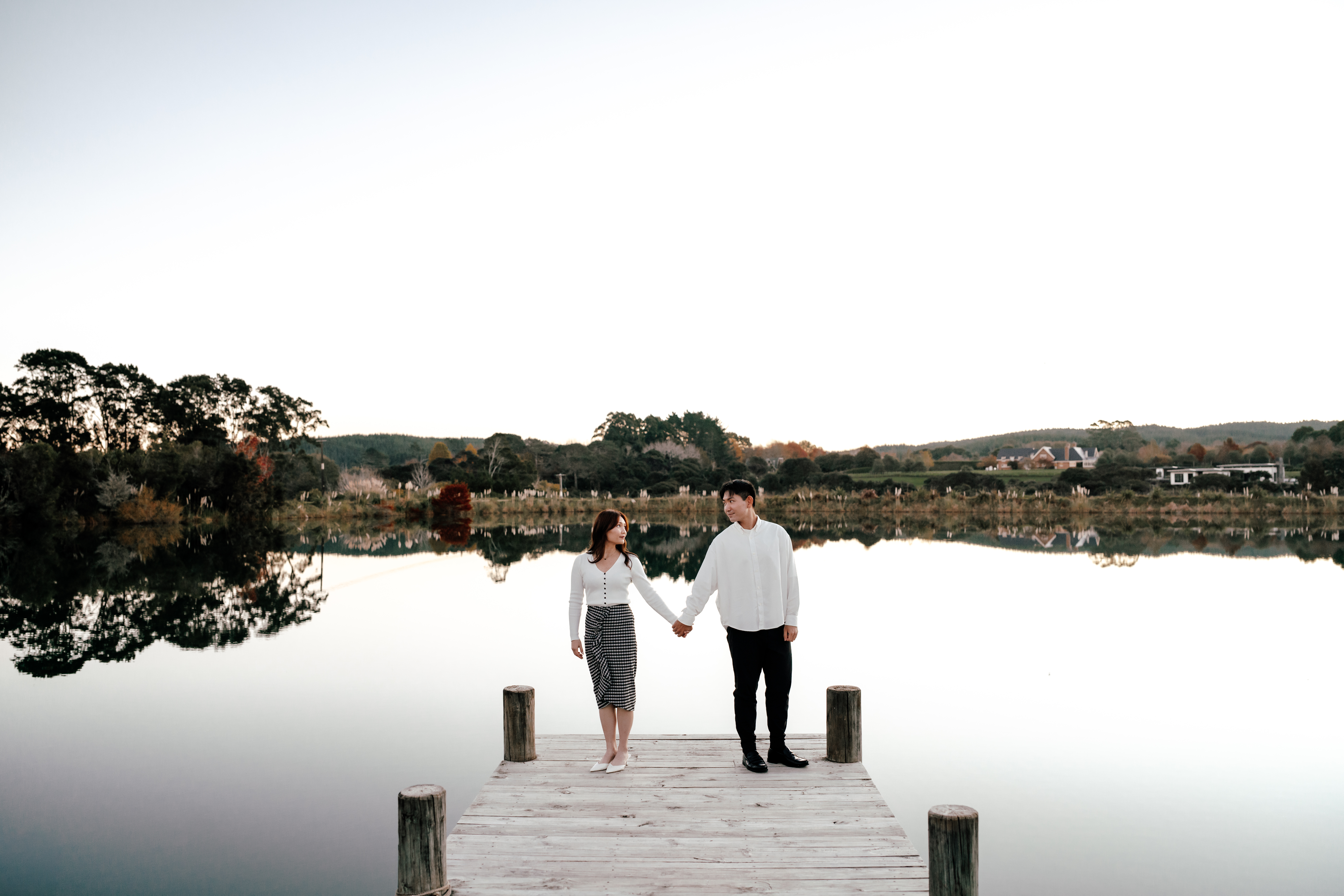 A couple standing hand in hand on a beautiful deck over a lake next to a house in Auckland during their proposal photoshoot by Zanthe Vorsatz Photography